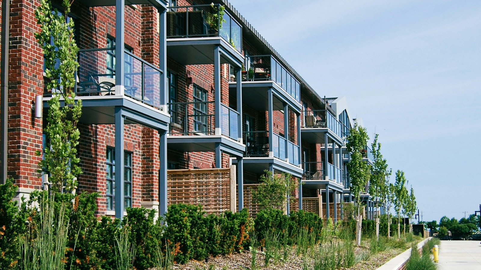 Low-rise red brick apartment building with glass balconies above landscaped garden beds and a walkway