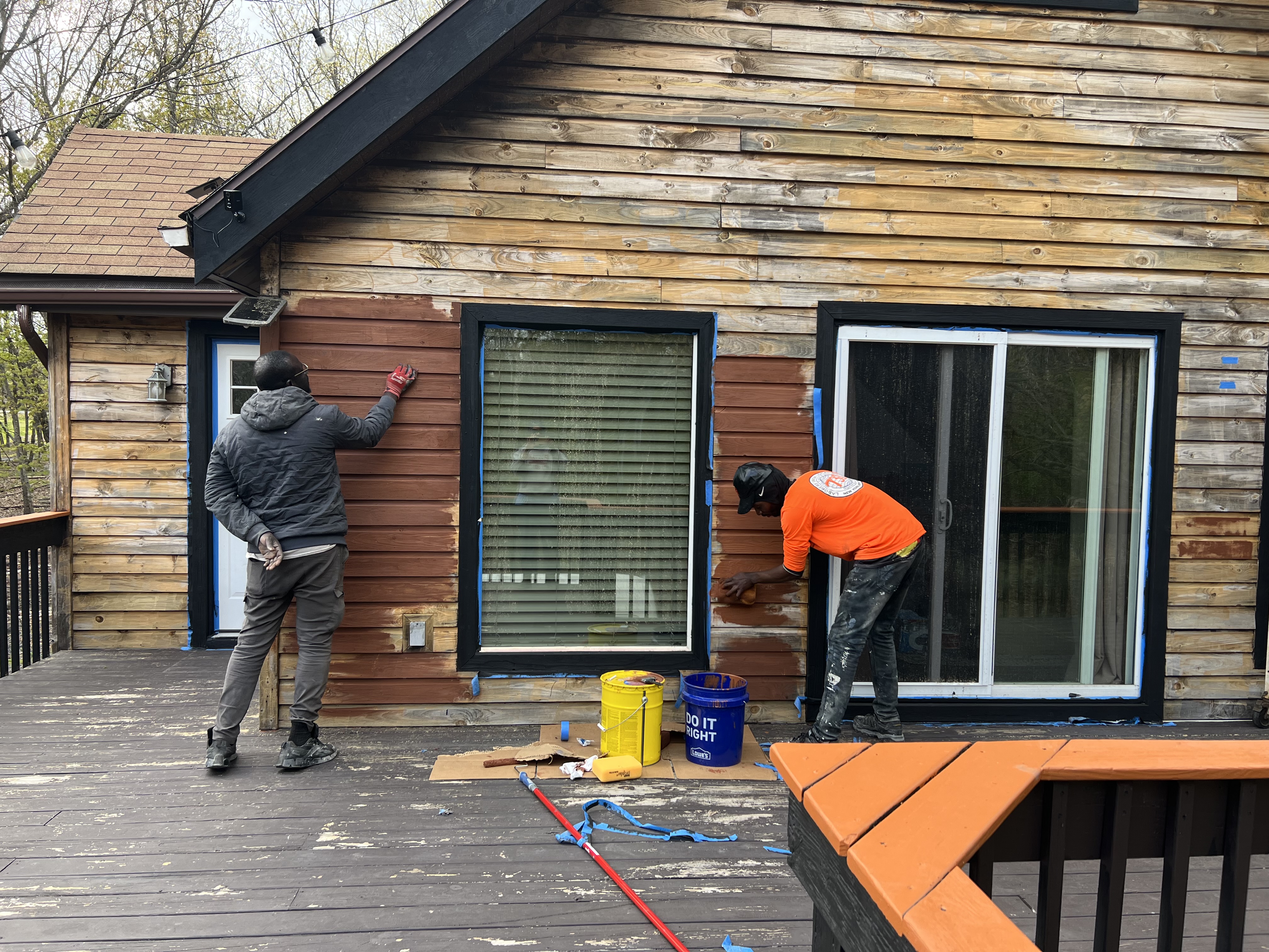 Poconos cabin in progress — two crew members staining weathered wood siding and deck wall