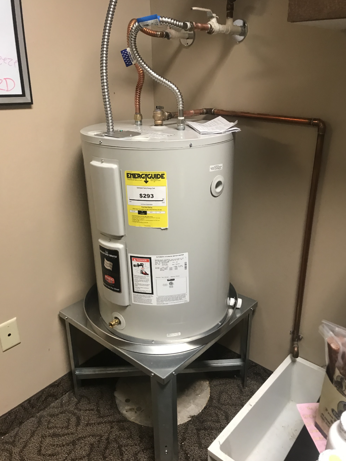 Bradford White electric water heater on a raised metal stand with drip pan and copper piping installed by Aurora Plumbing & Mechanical
