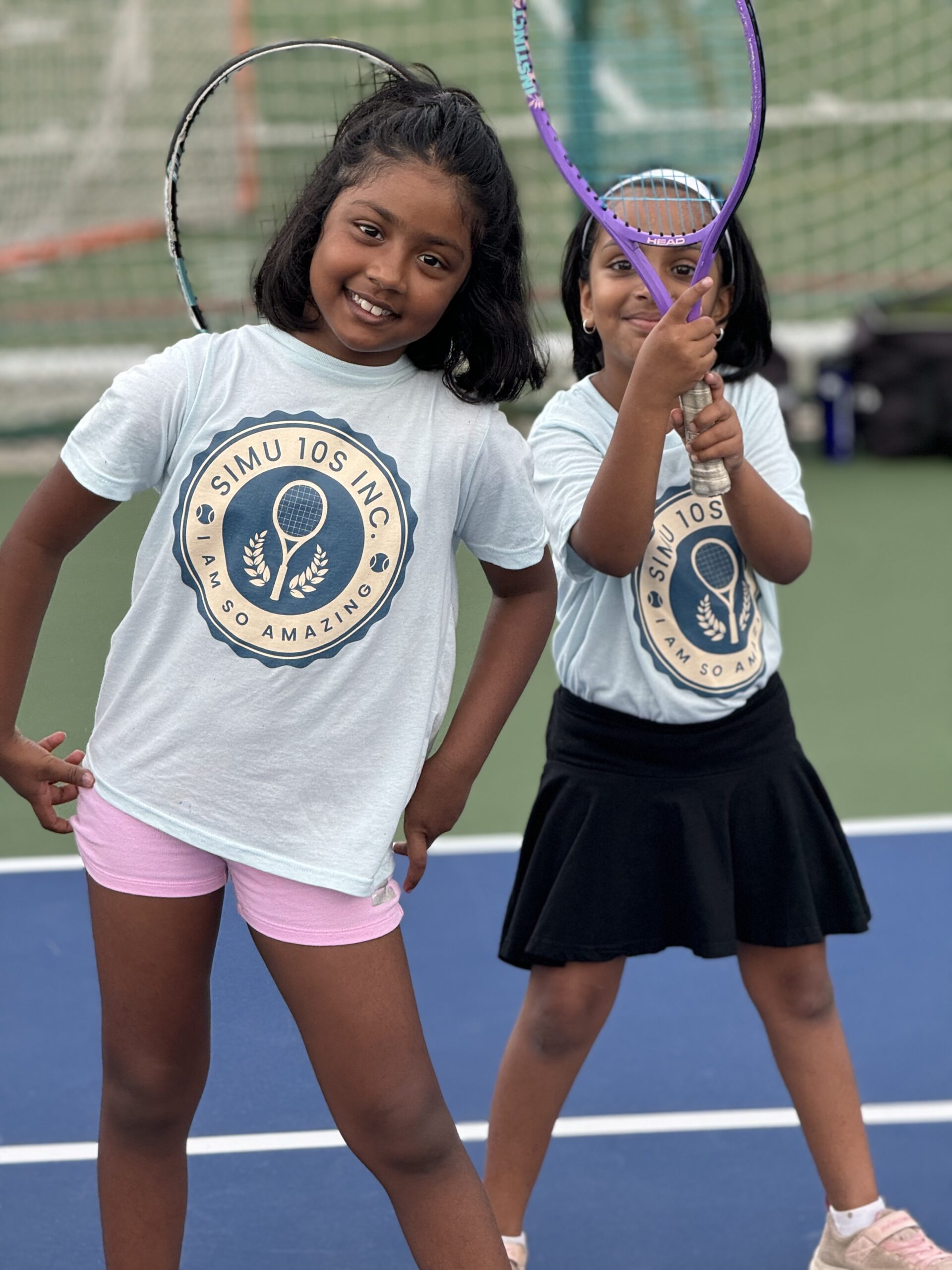 Two young Simu 10s players smiling and holding tennis rackets courtside