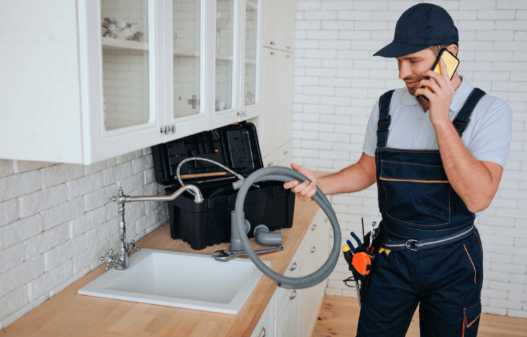 Plumber working on sink pipe and answering phone in kitchen