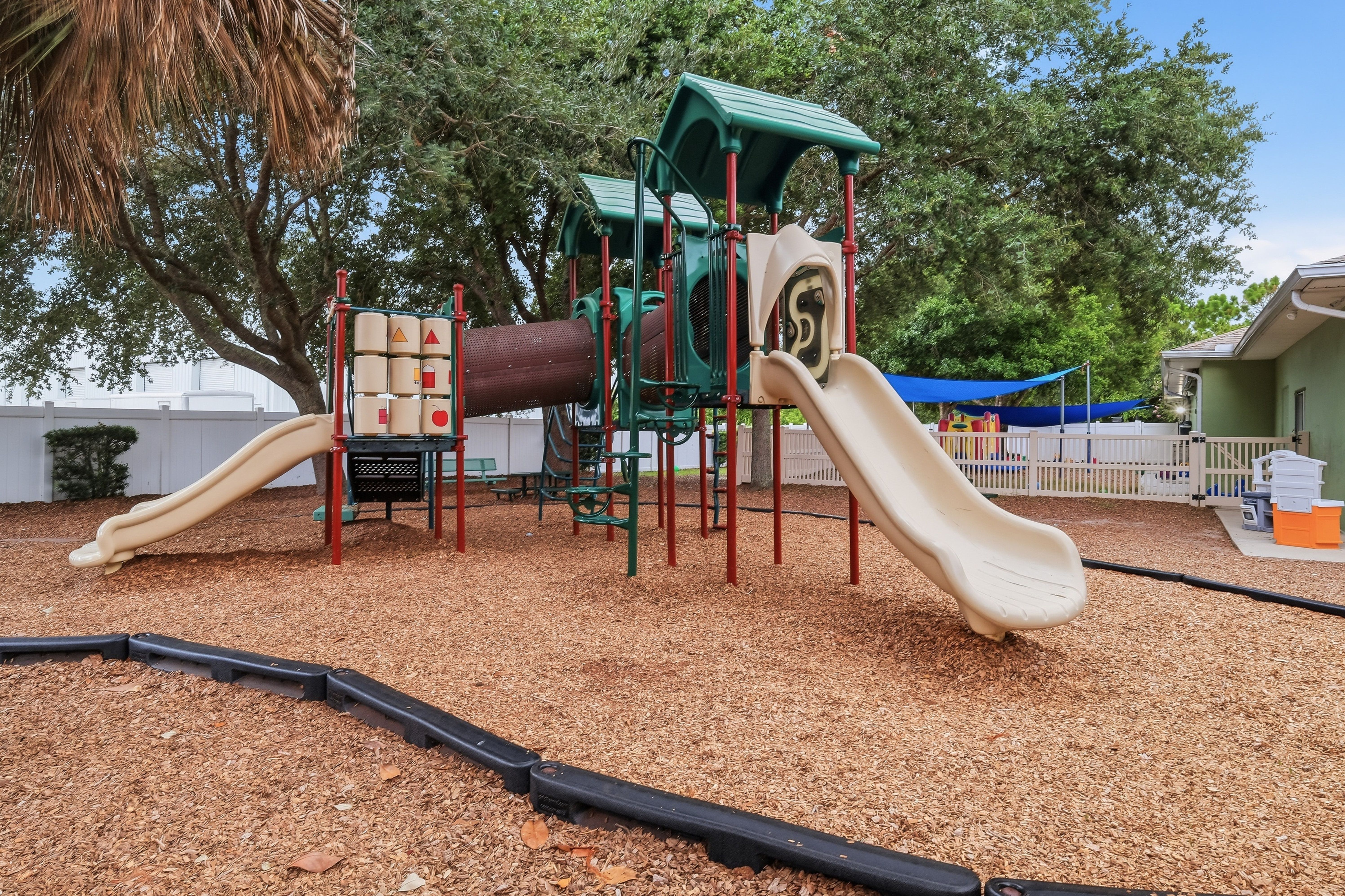 Outdoor playground with multi-level play structure, slides, and wood chip mulch under mature shade trees