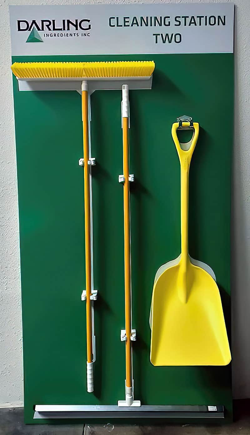 Darling Ingredients Cleaning Station Two deep green shadow board with high-visibility yellow push broom, floor squeegee, and scoop shovel
