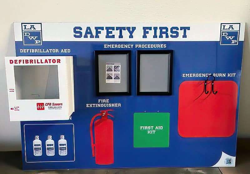 LADWP Safety First emergency visual center with Defibrillator AED cabinet, emergency procedures snap frames, fire extinguisher silhouette, eyewash bottles, first aid kit, and emergency burn kit
