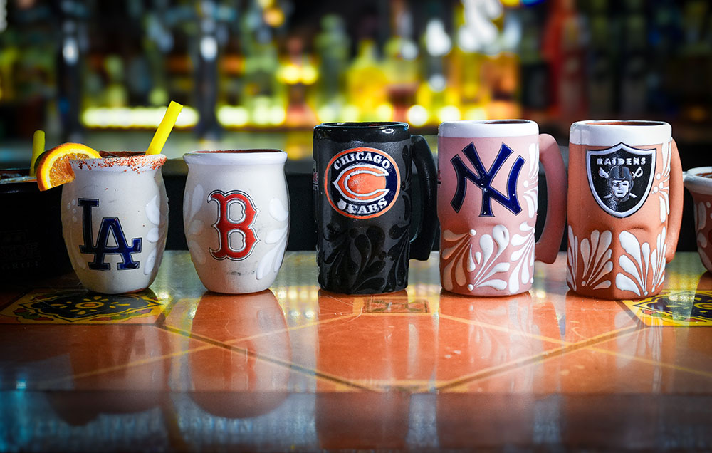 Custom sports team mugs lined up on the bar at Casa Grande Stellhorn Road featuring Dodgers, Red Sox, Bears, Yankees, and Raiders designs with festive Mexican floral patterns
