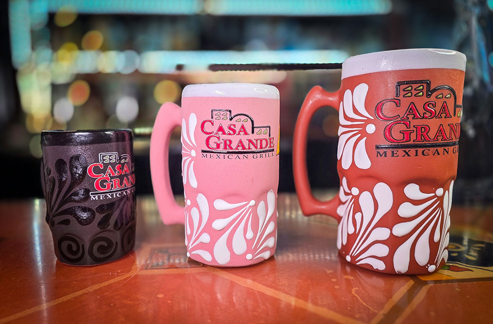 Three sizes of Casa Grande custom ceramic mugs showing black, pink, and terracotta clay finishes with white floral relief
