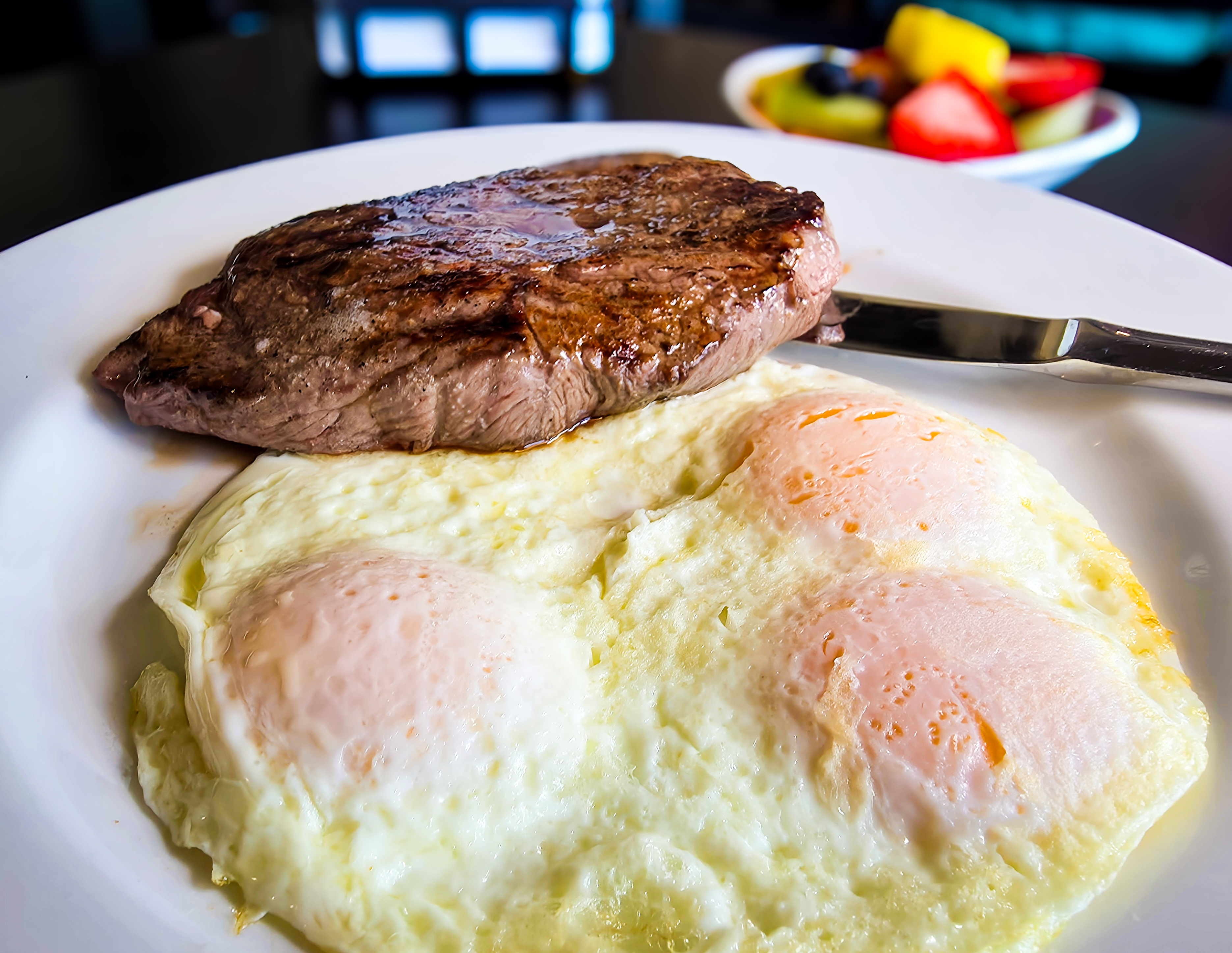 Juicy seared steak paired with three sunny-side eggs and fresh fruit bowl