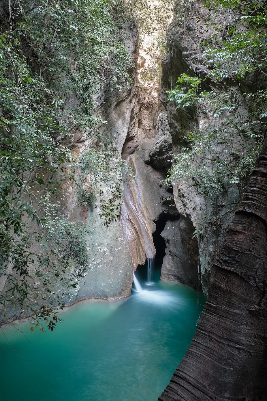 Dramatic vertical rock canyon with a narrow mountain spring waterfall spilling into a deep emerald pool sheltered by jungle trees in Samboan, Southern Cebu