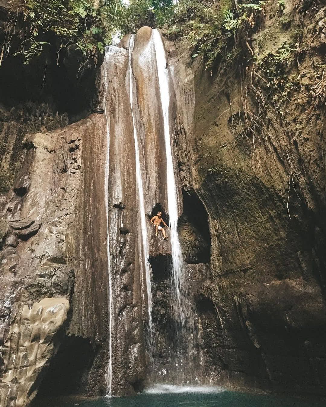 Swimmer sitting in the natural rock alcove of Binalayan Hidden Falls in Samboan, Southern Cebu, framed by towering canyon walls and plunging cascade