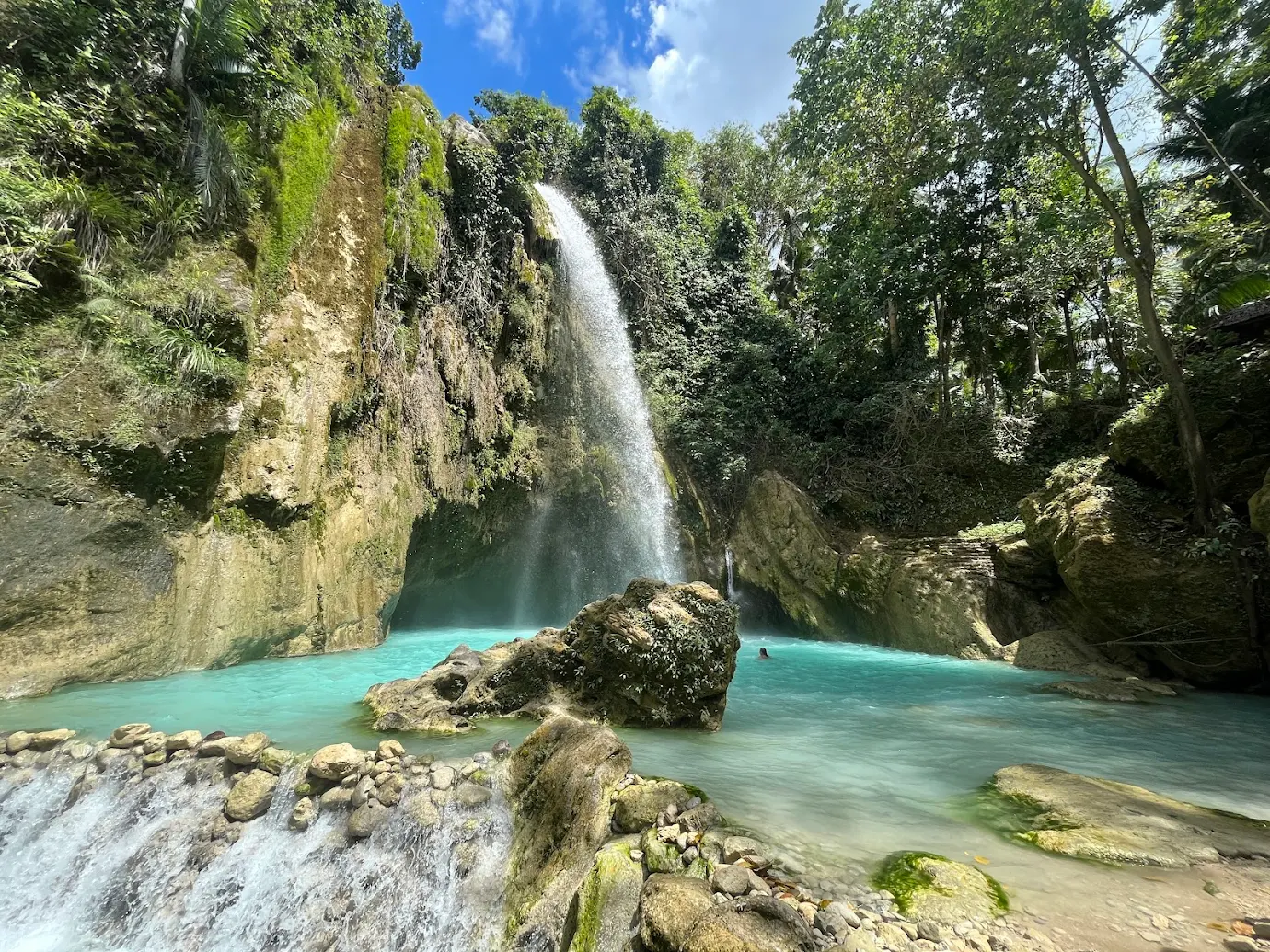 Sunlit tropical waterfall pouring into a wide clear cyan pool with a swimmer enjoying the fresh mountain water in Samboan, Southern Cebu
