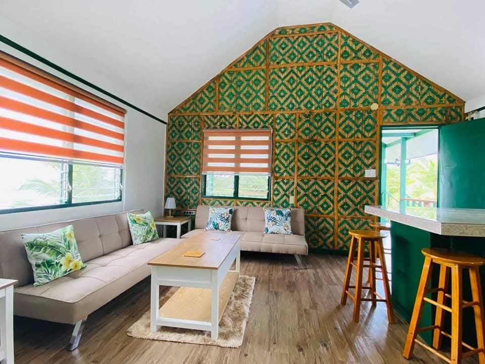 High-ceiling living lounge at Mims House Cebu featuring woven geometric amakan bamboo accent wall, comfortable futon sofas, coffee table, and kitchen breakfast bar with wooden stools