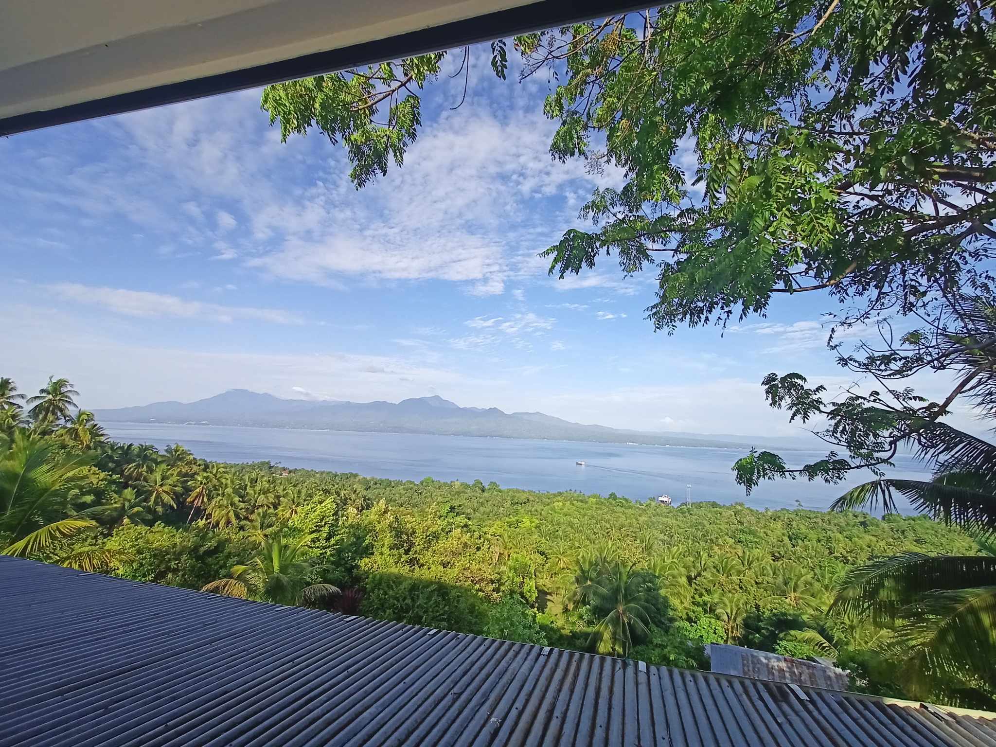 High elevation panorama framed above the corrugated rooftop and lush green treetops looking across the strait to distant Negros mountain peaks and a boat