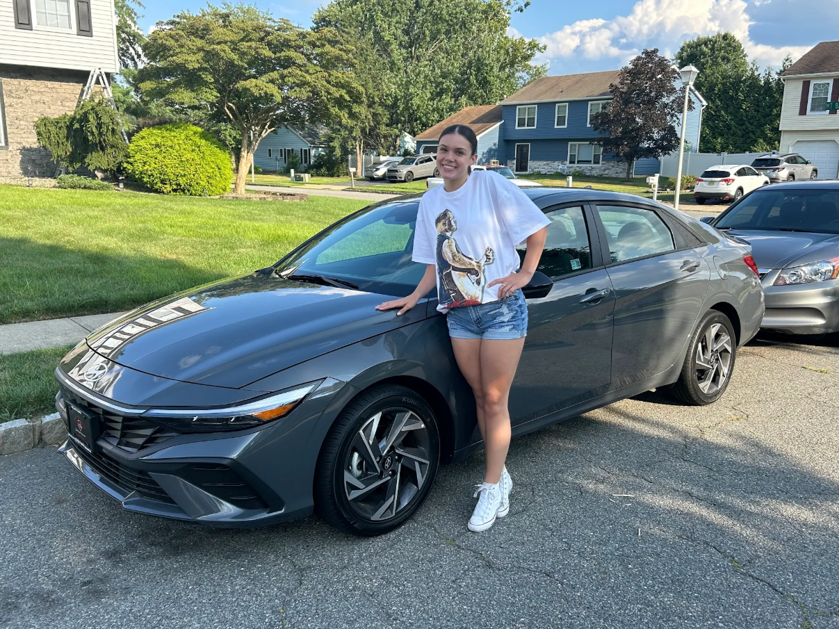 Customer smiling alongside newly delivered gray sedan parked on a residential street