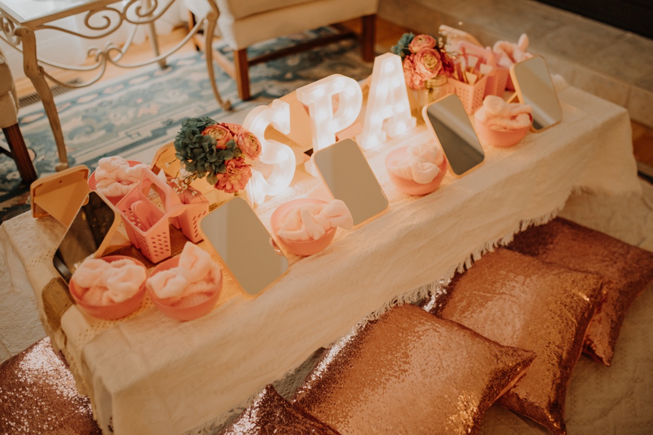 Full styled spa party setup with marquee light-up SPA letters, rose gold sequin floor cushions, individual mirrors, bowls, and pink headbands