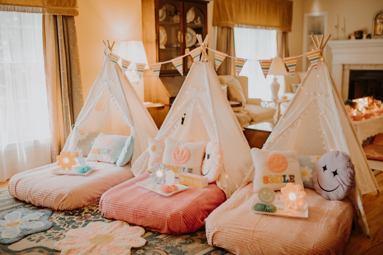Three white canvas teepees with colorful pennant bunting, daisy floor rugs, and glowing fairy lights