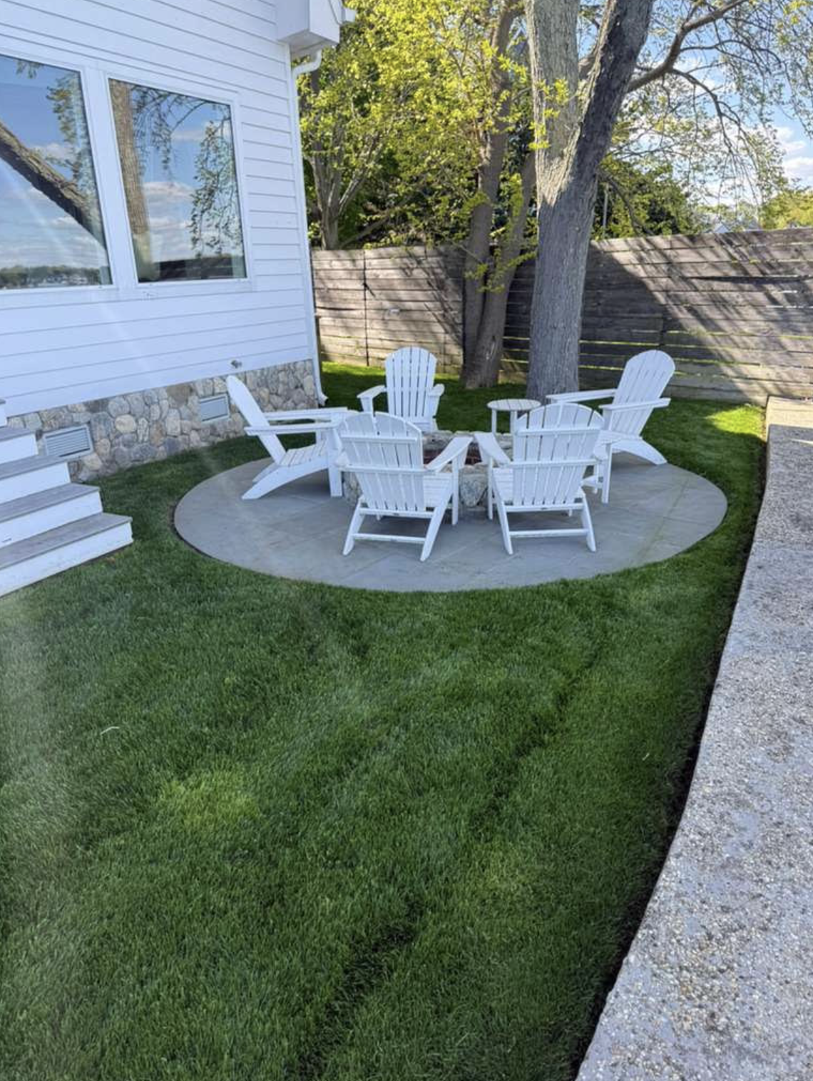 Completed backyard outdoor living space: custom circular stone fire pit patio with white Adirondack seating and lush grass