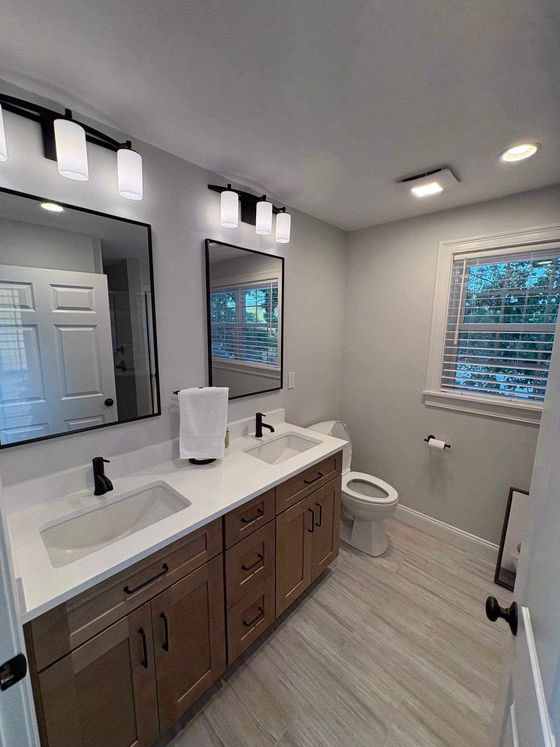 Completed master bathroom renovation with warm wood double vanity, black modern hardware, and bright recessed lighting
