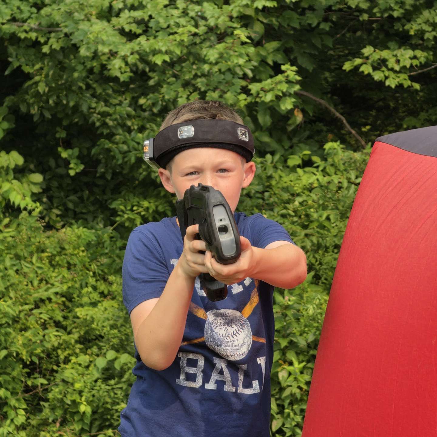 Young camper wearing sensor headband taking aim beside a red inflatable bunker during camp laser tag