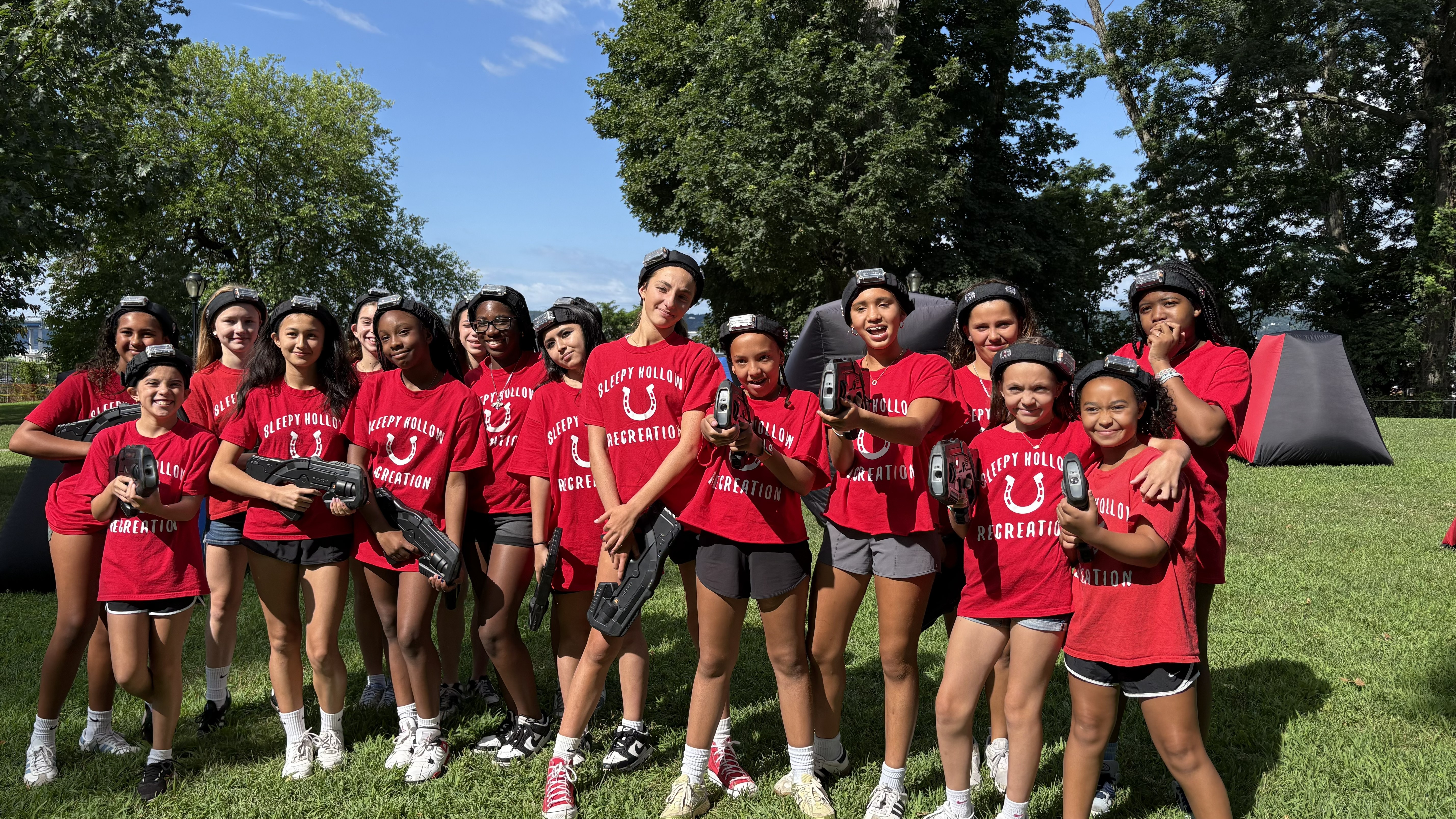 Recreation summer camp group of girls holding laser tag blasters and wearing sensor headbands outdoors with inflatable bunkers