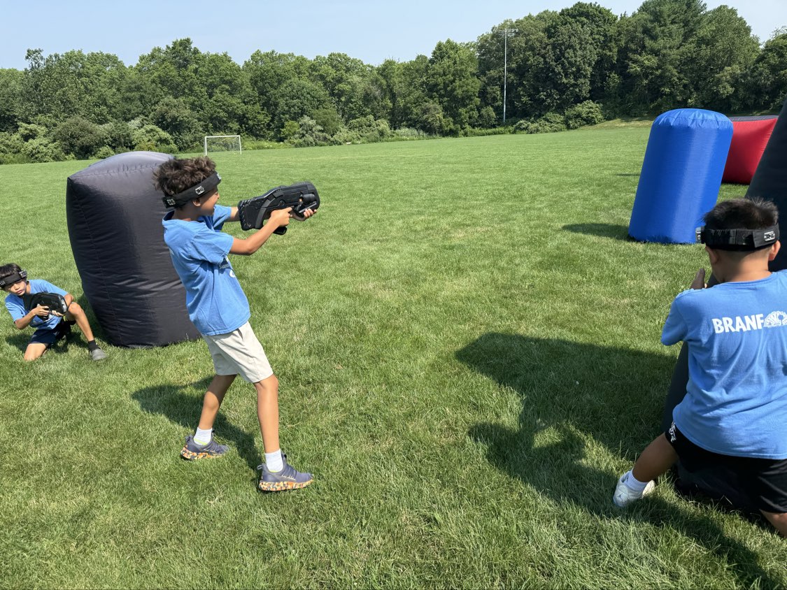 Summer camp campers in blue shirts playing mobile laser tag on an open field with inflatable bunkers