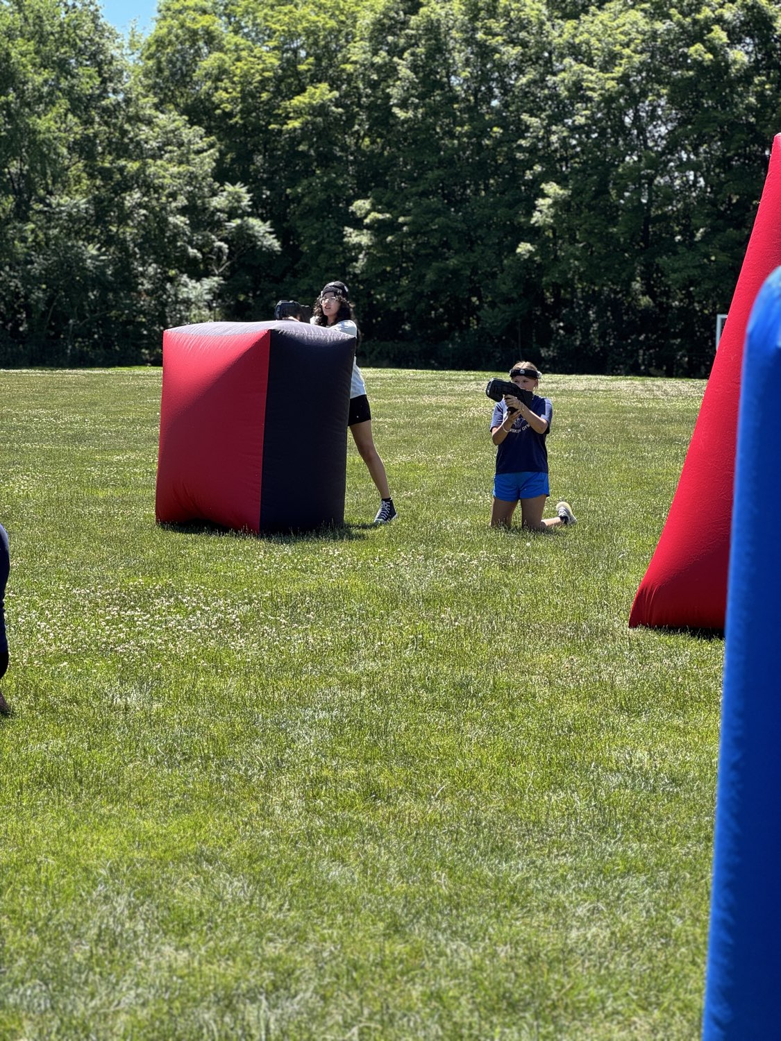 Campers maneuvering and aiming from behind red and blue inflatable bunkers on a wide summer camp field
