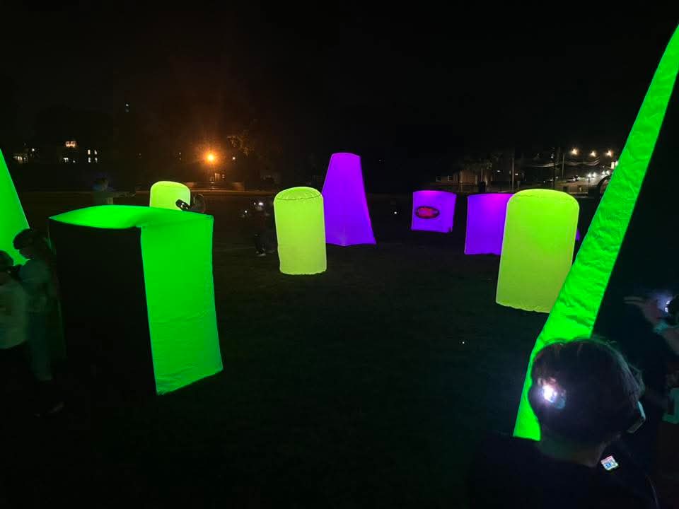 Nighttime outdoor laser tag event with green and purple illuminated inflatable bunkers