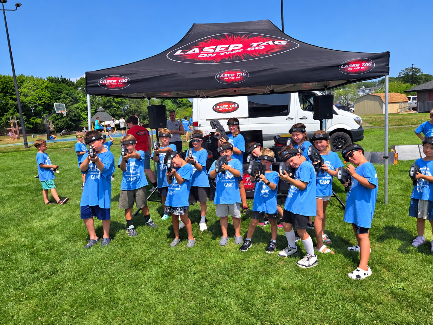 Summer camp kids in blue camp shirts posing with blasters and headbands under the Laser Tag On The Go branded event tent with mobile van