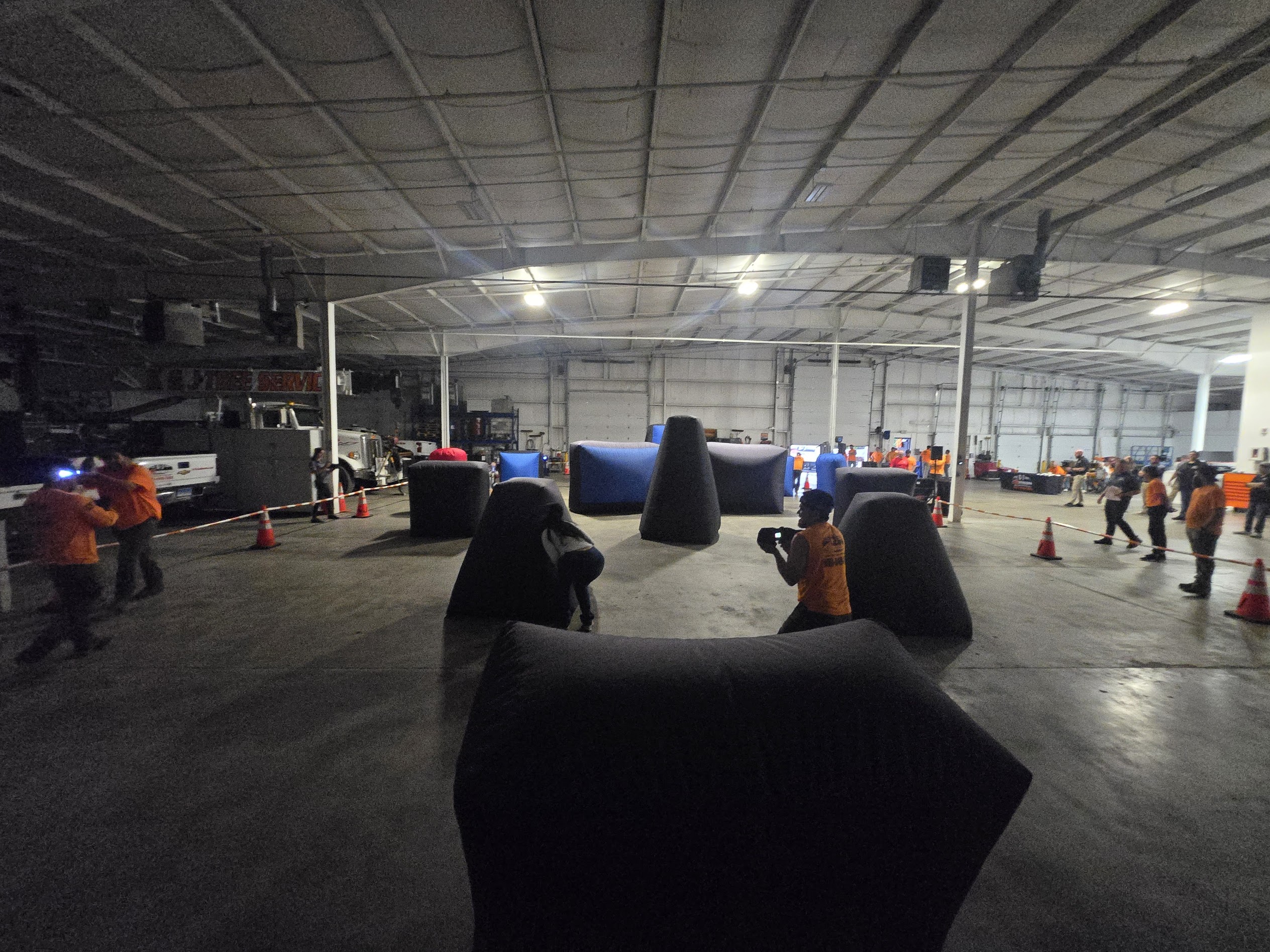 Corporate laser tag event in a wide indoor warehouse arena with players in orange shirts among black and blue inflatable bunkers