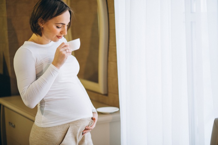 Mujer embarazada de pie junto a la ventana bebiendo una infusión suave