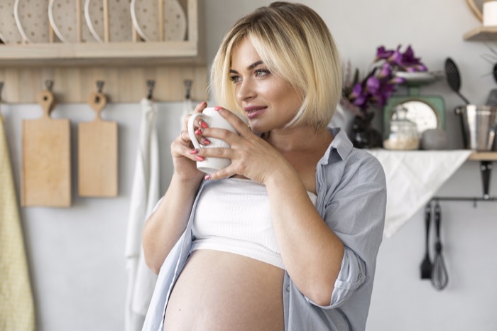 Mujer embarazada sonriendo en la cocina sosteniendo una taza de infusión tibia