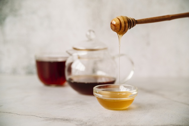 Cuchara de madera vertiendo miel natural junto a tetera y taza de té caliente