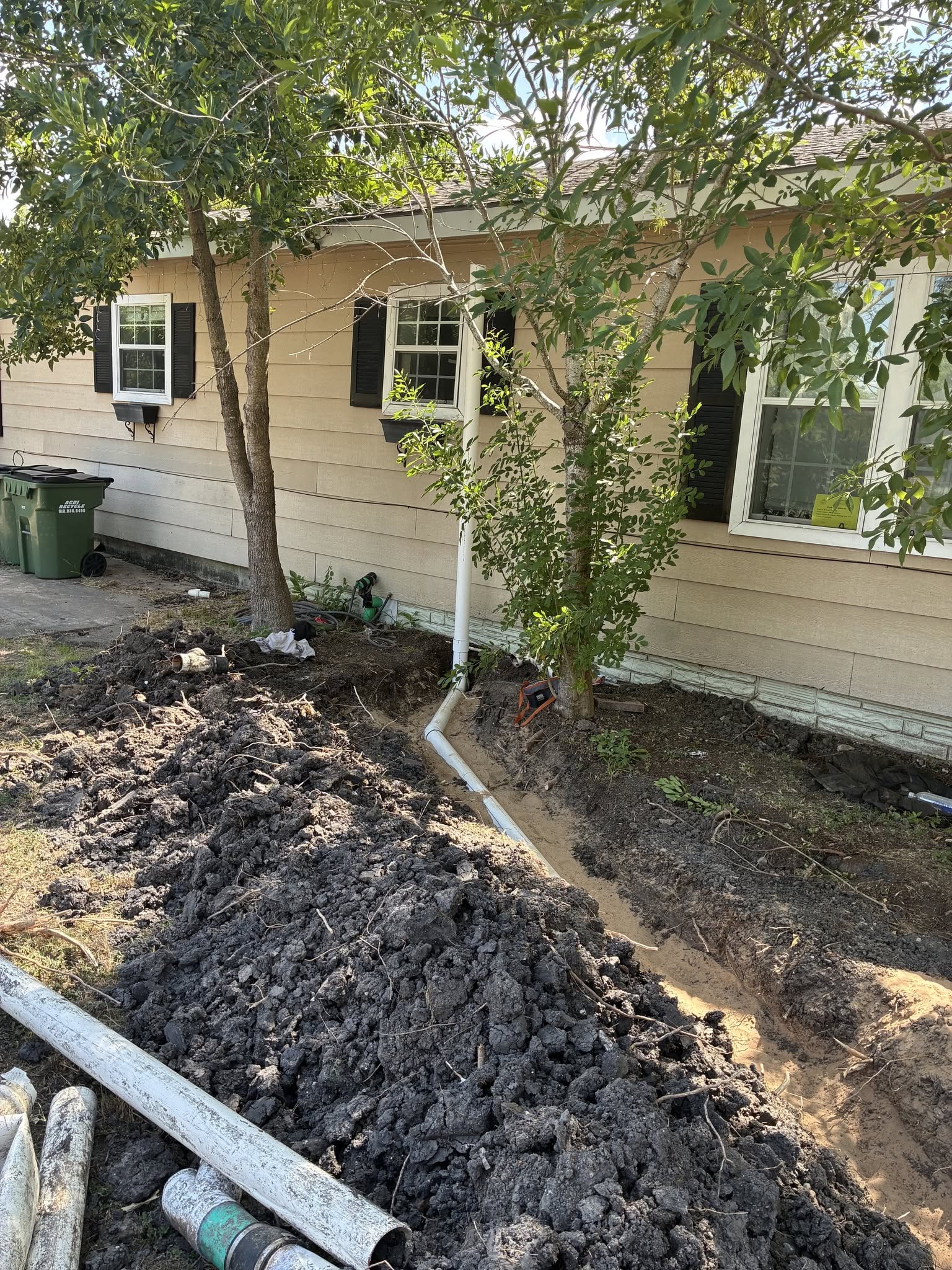 Excavated sewer trench along residential home foundation showing new PVC line bedded in sand with stack tie-in