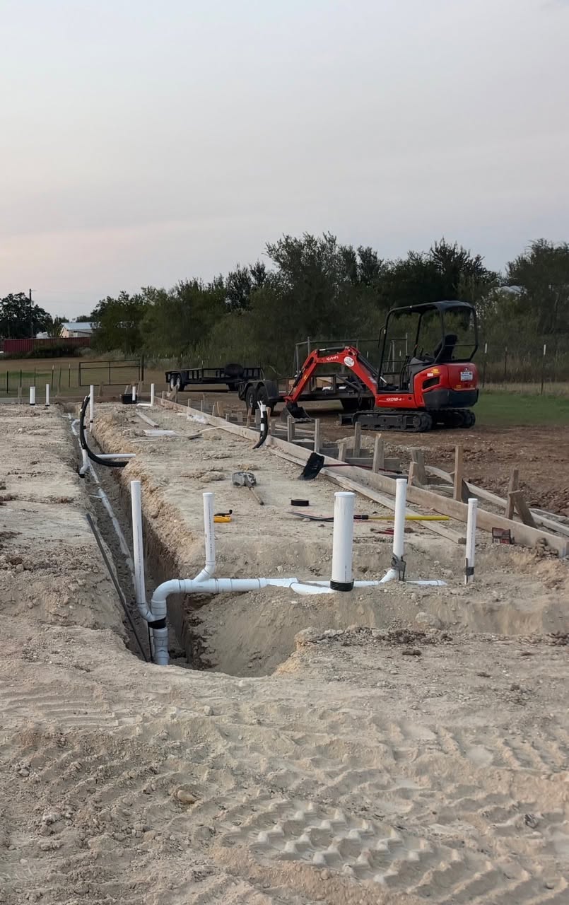 Underground plumbing rough-in trench with cleanouts, drain lines, and sleeves positioned before concrete pour