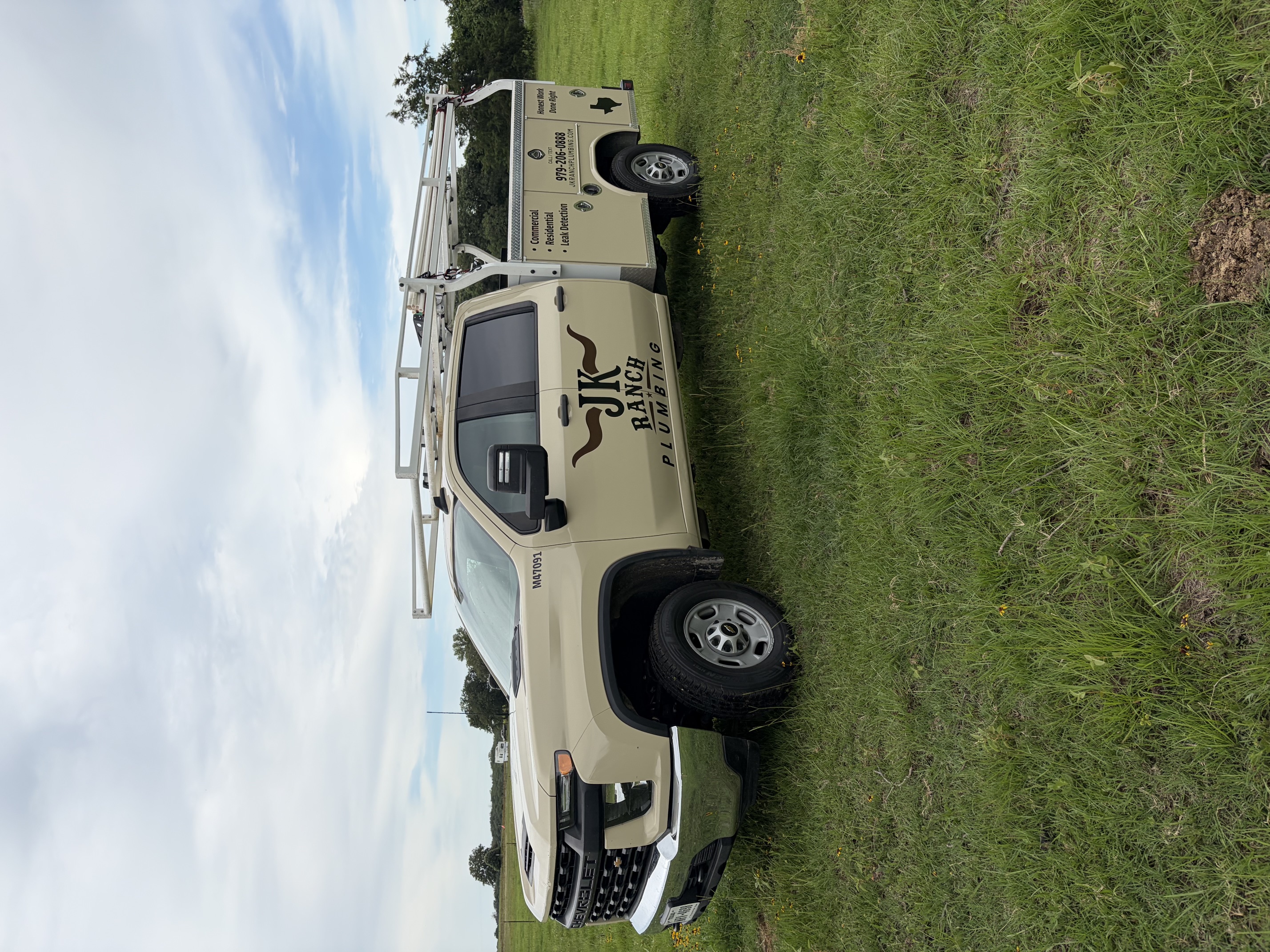 Side profile of the cream JK Ranch Plumbing truck in the field showing logo, phone number, and service body