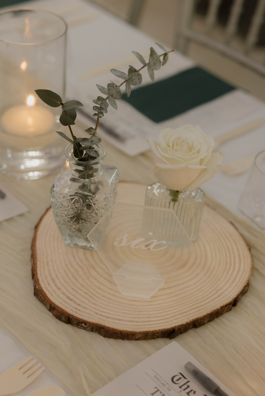 Wood slice centerpiece with delicate bud vases, single ivory rose, seeded eucalyptus, glowing candlelight, and custom acrylic table number six