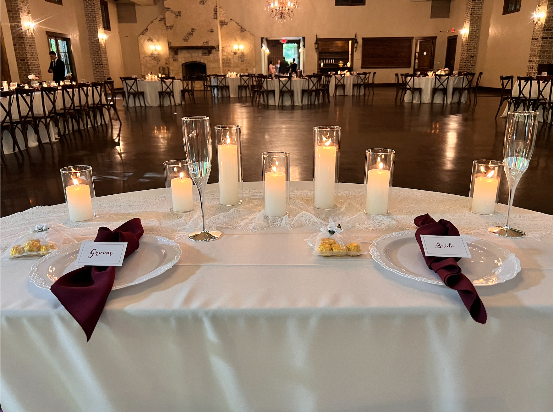 Grand ballroom reception venue styled by HoneyBee Events with a candlelit sweetheart table, burgundy napkins, personalized bride and groom place settings, and illuminated guest tables surrounding the dance floor