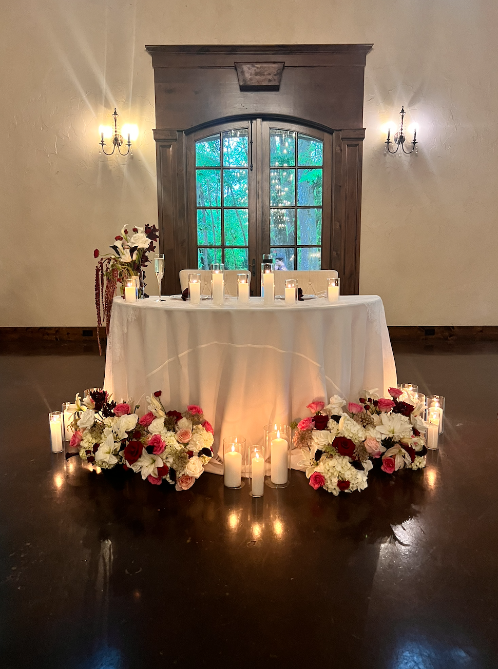 Candlelit sweetheart table styled with soft ivory linen, glowing hurricane pillar candles, and a lush floral meadow of roses and hydrangeas