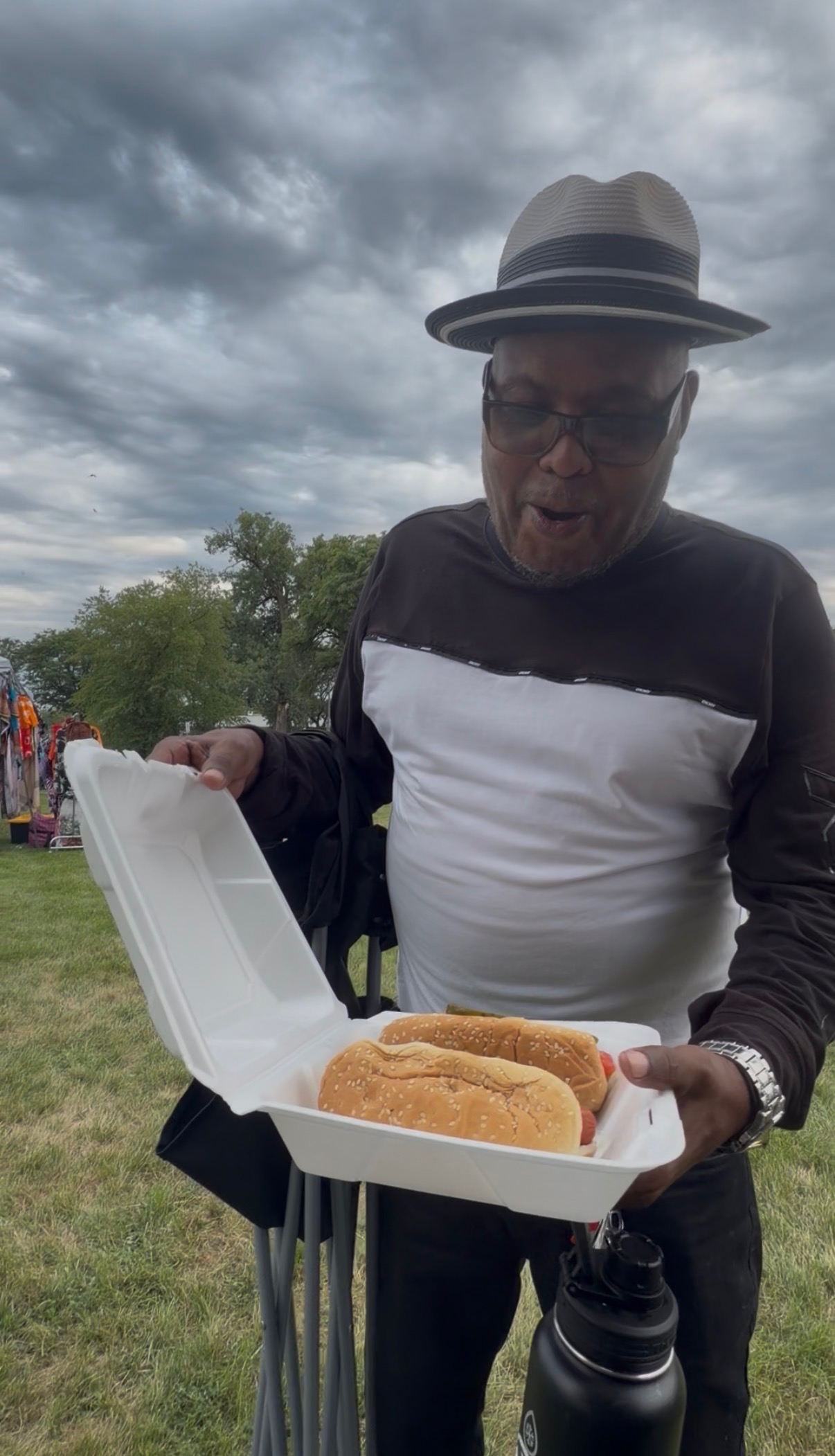 Customer under a cloudy sky opening a takeout box with two loaded hot dogs