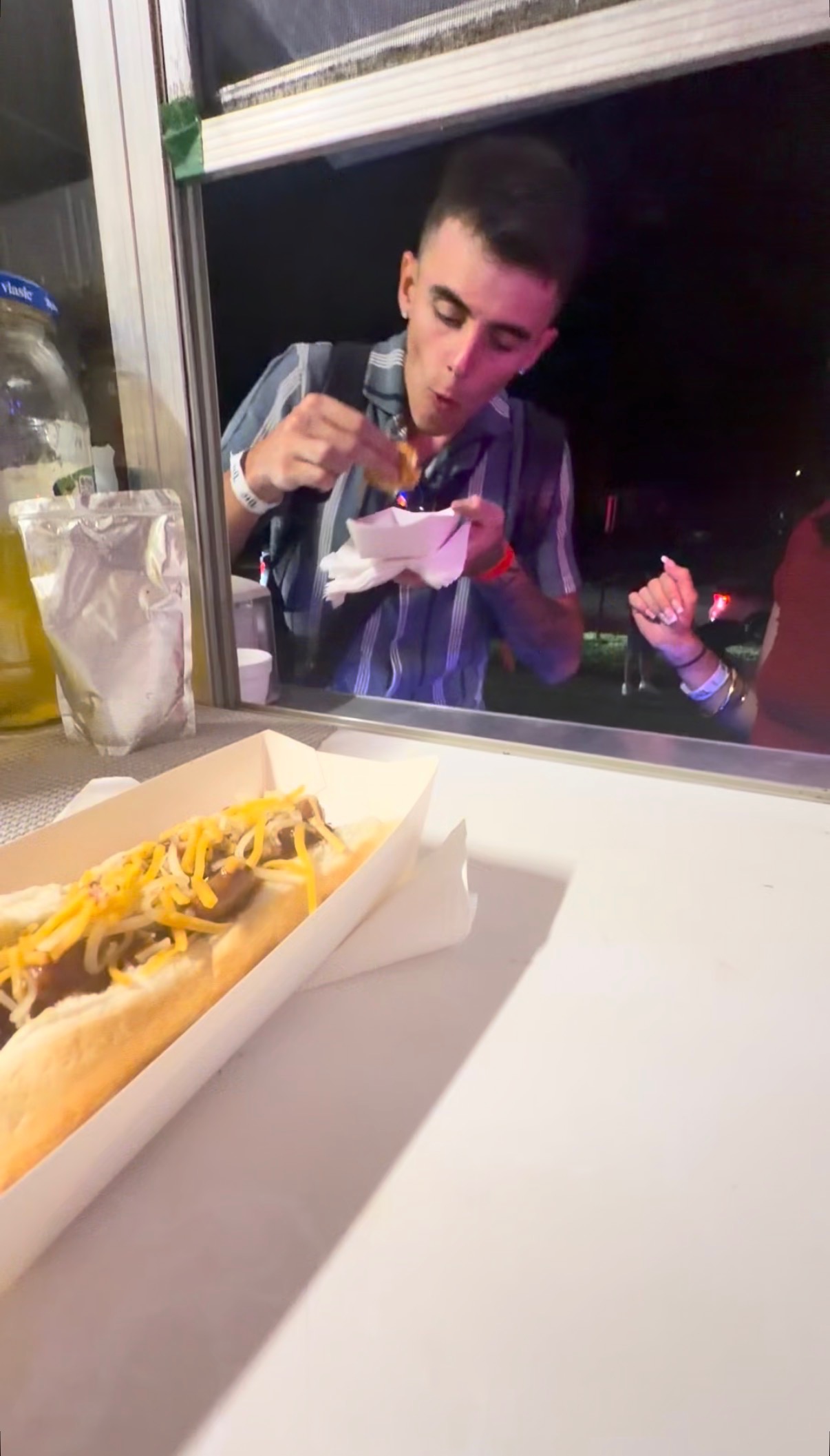 Customer at the Papa Dogs trailer window enjoying a loaded chili cheese dog at night