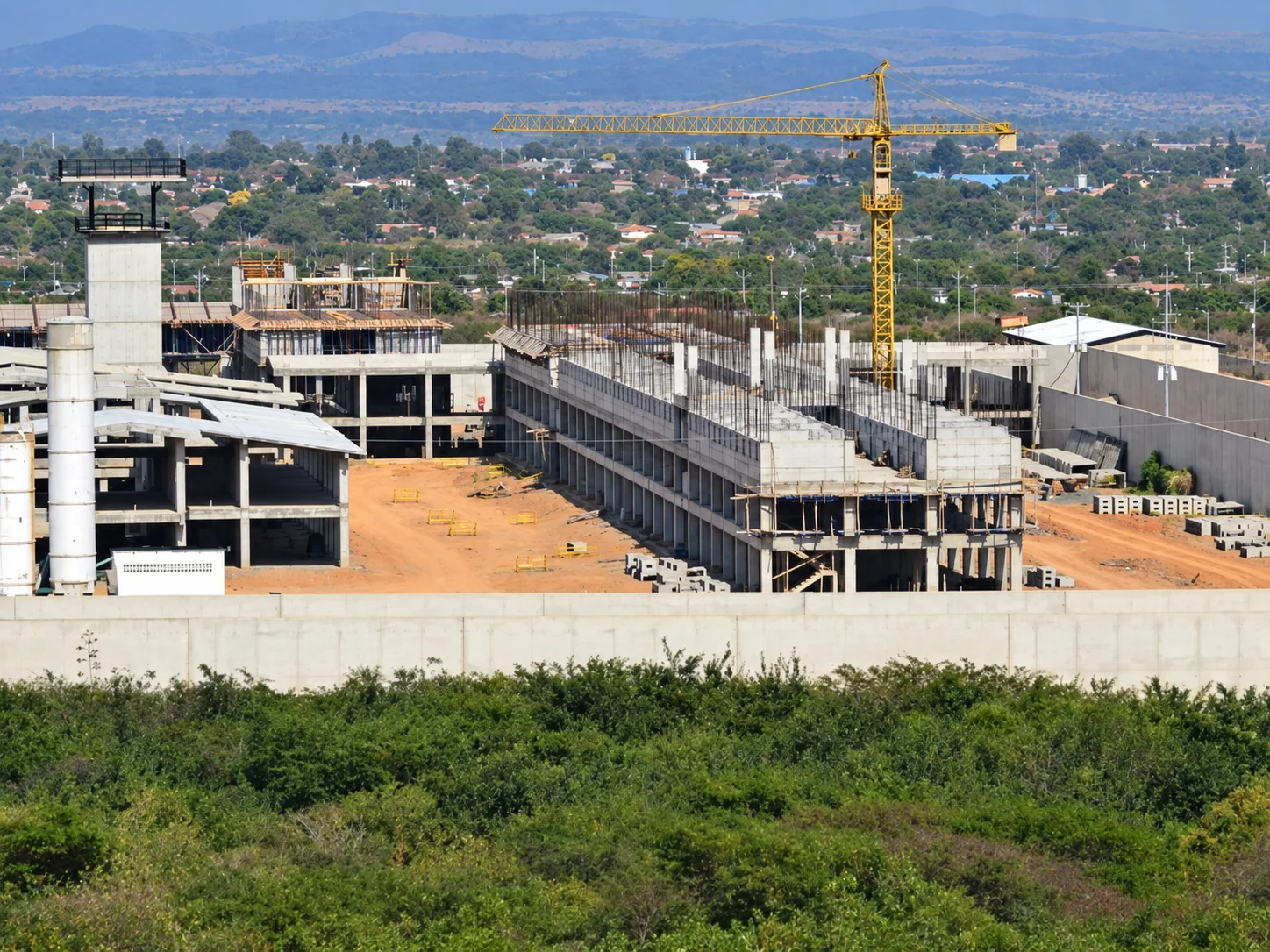 Fase de construcción estructural de pabellones con grúa torre, torre de control y vigilancia, y muros perimetrales de contención