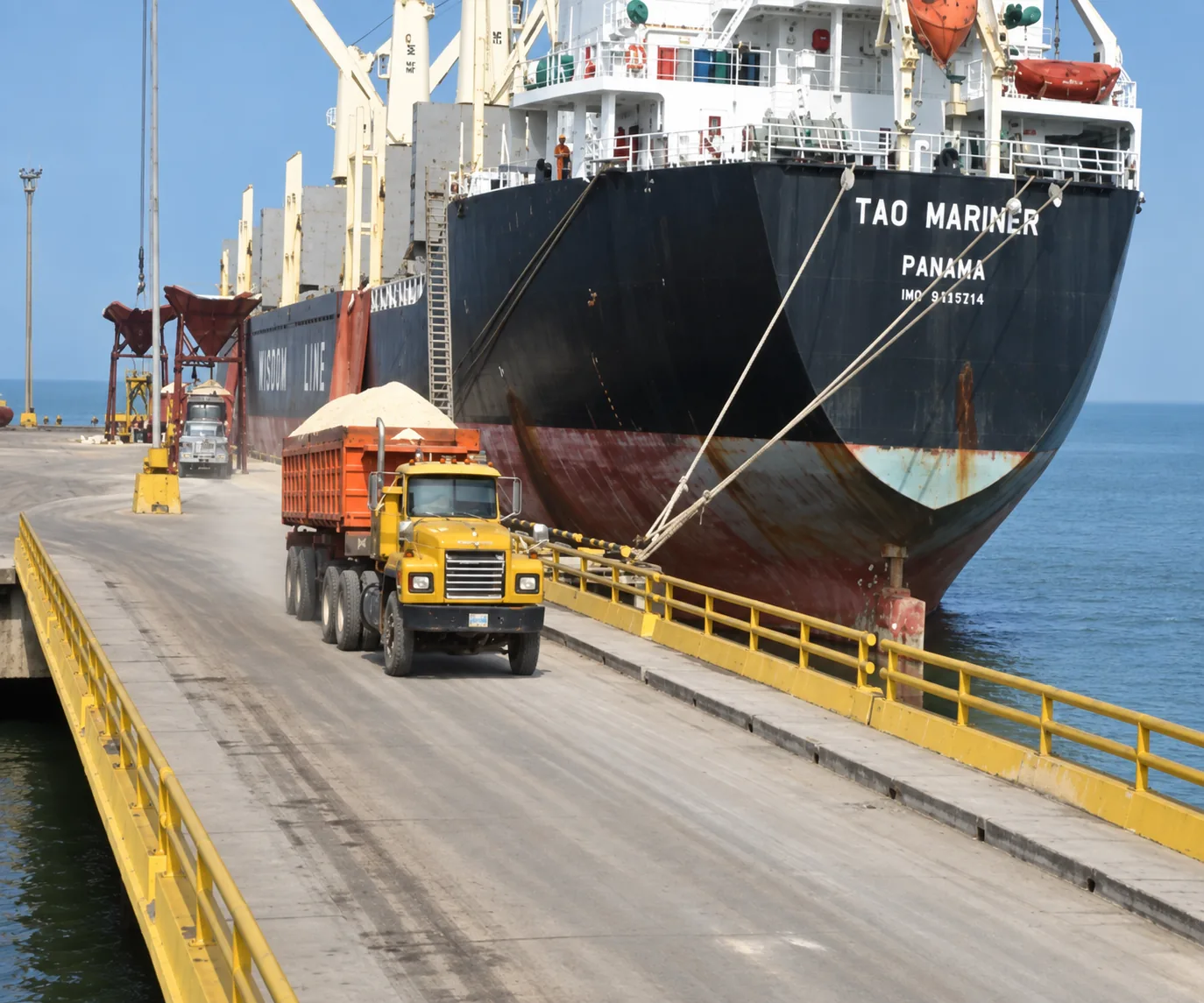 Operación de carga y descarga de buque granelero acoderado al muelle con tránsito continuo de camiones de carga pesada por el viaducto
