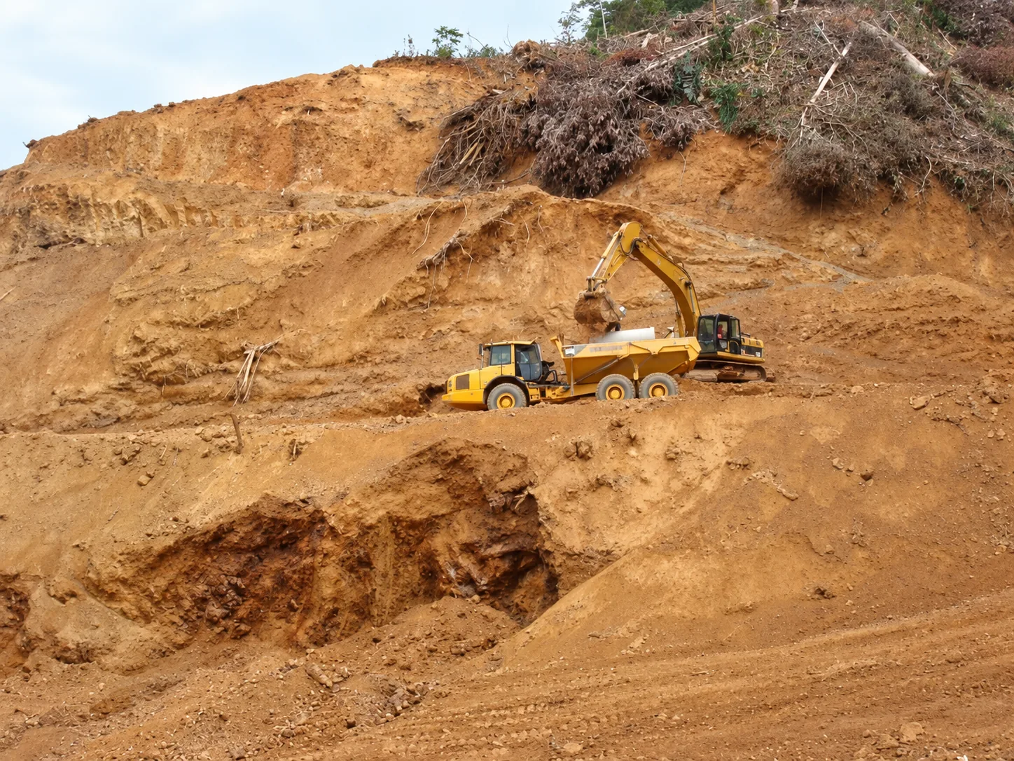 Cortes de ladera, banqueo y conformación de terrazas del sitio con retroexcavadora de orugas cargando camión articulado todoterreno