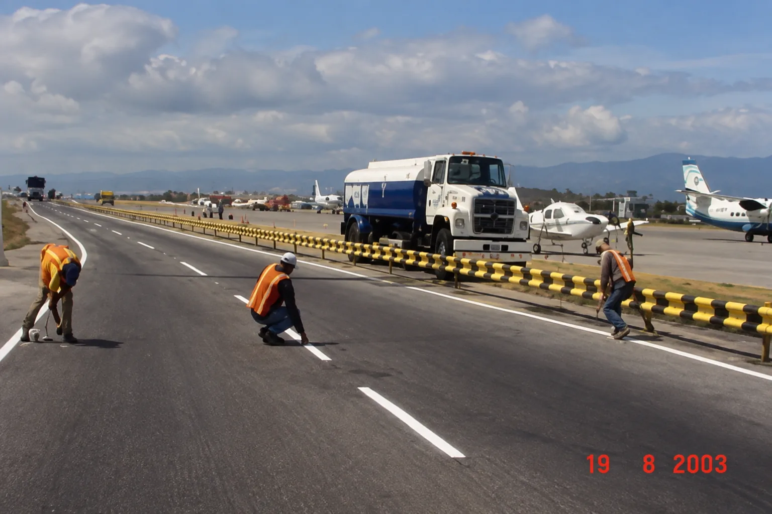 Cuadrilla técnica ejecutando labores de demarcación vial y señalización horizontal sobre la vía interna de servicio con camión cisterna de soporte operativo y aeronaves al fondo (agosto 2003)