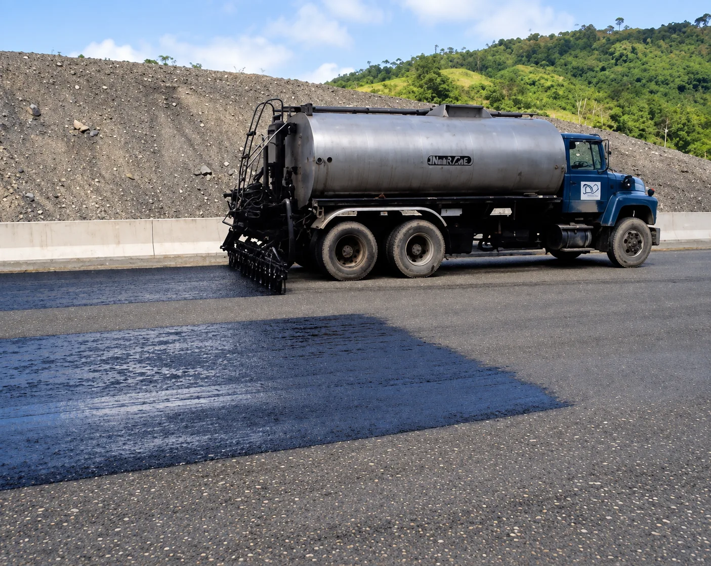 Camión distribuidor de asfalto aplicando riego de imprimación bituminosa mediante barra regadora a presión sobre la base granular del pavimento