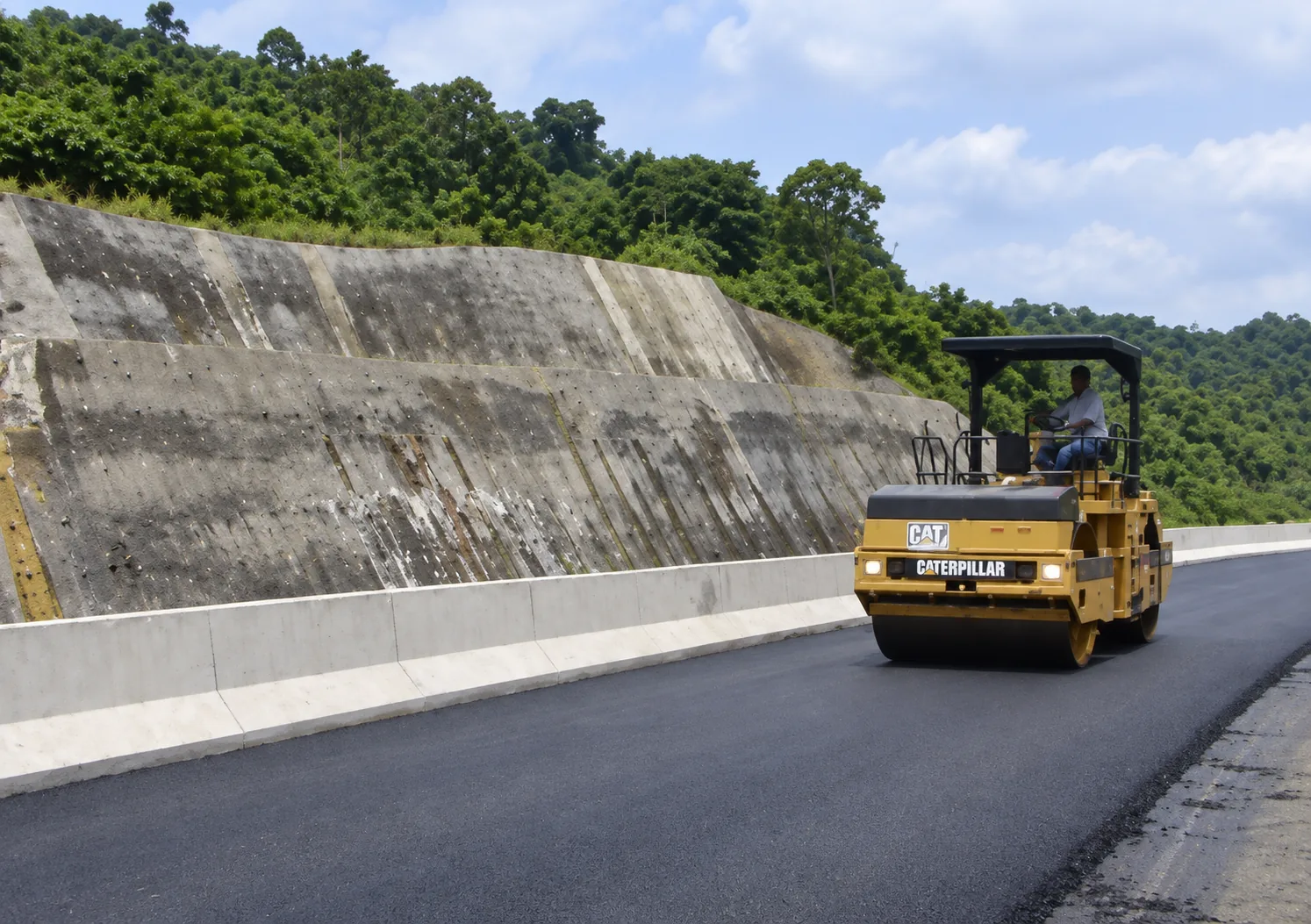 Compactación de rodadura asfáltica con rodillo tándem vibratorio Caterpillar junto a barrera New Jersey y pantalla de sostenimiento