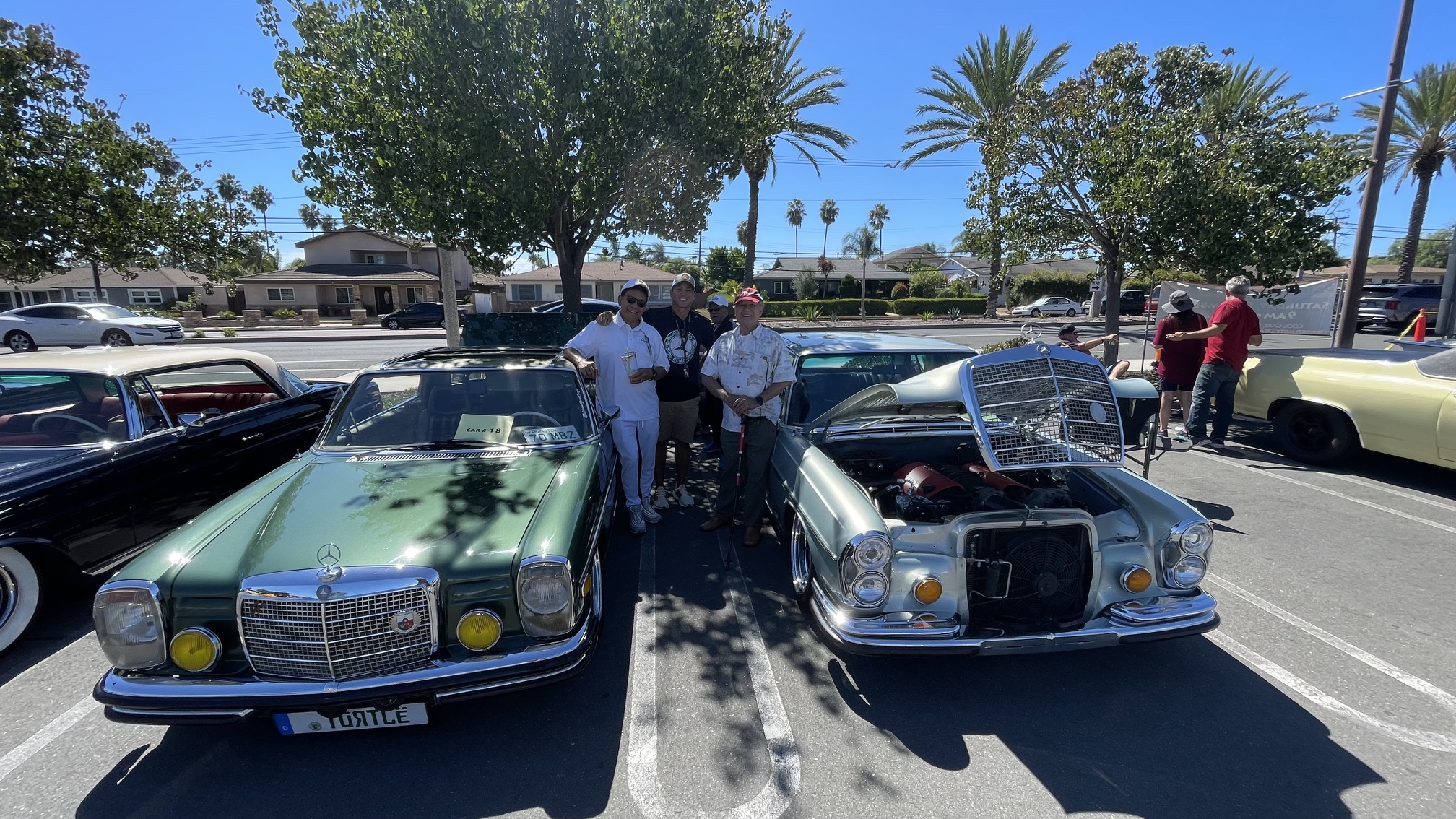 Parish car show enthusiasts smiling together between vintage green Mercedes-Benz and silver classic sedan