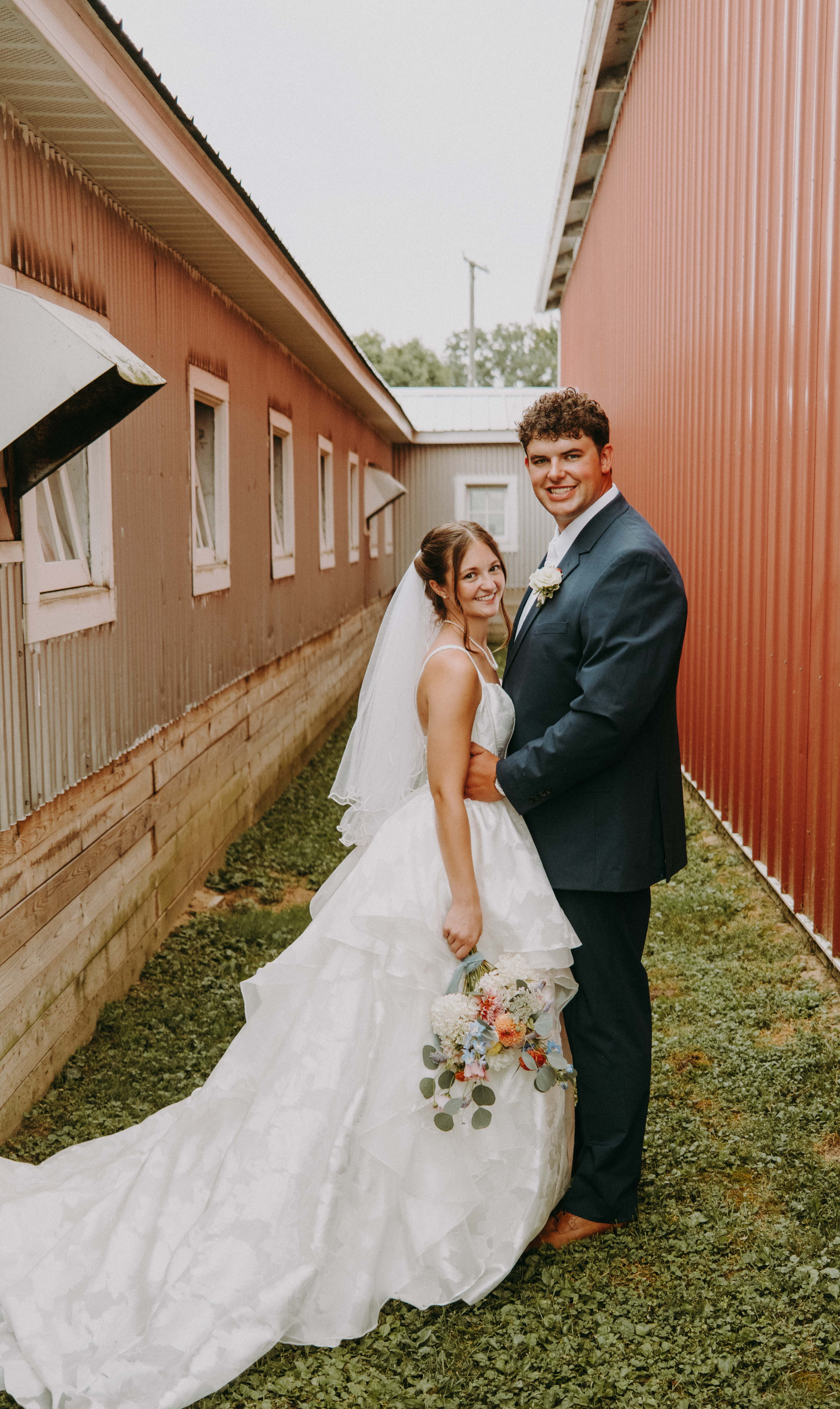 Smiling bride and groom standing outside barn with bridal bouquet