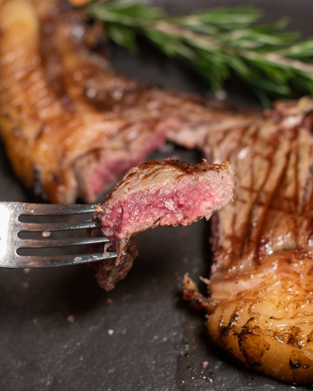 Boccone di carne cotta alla perfezione su forchetta con costata e rosmarino sullo sfondo