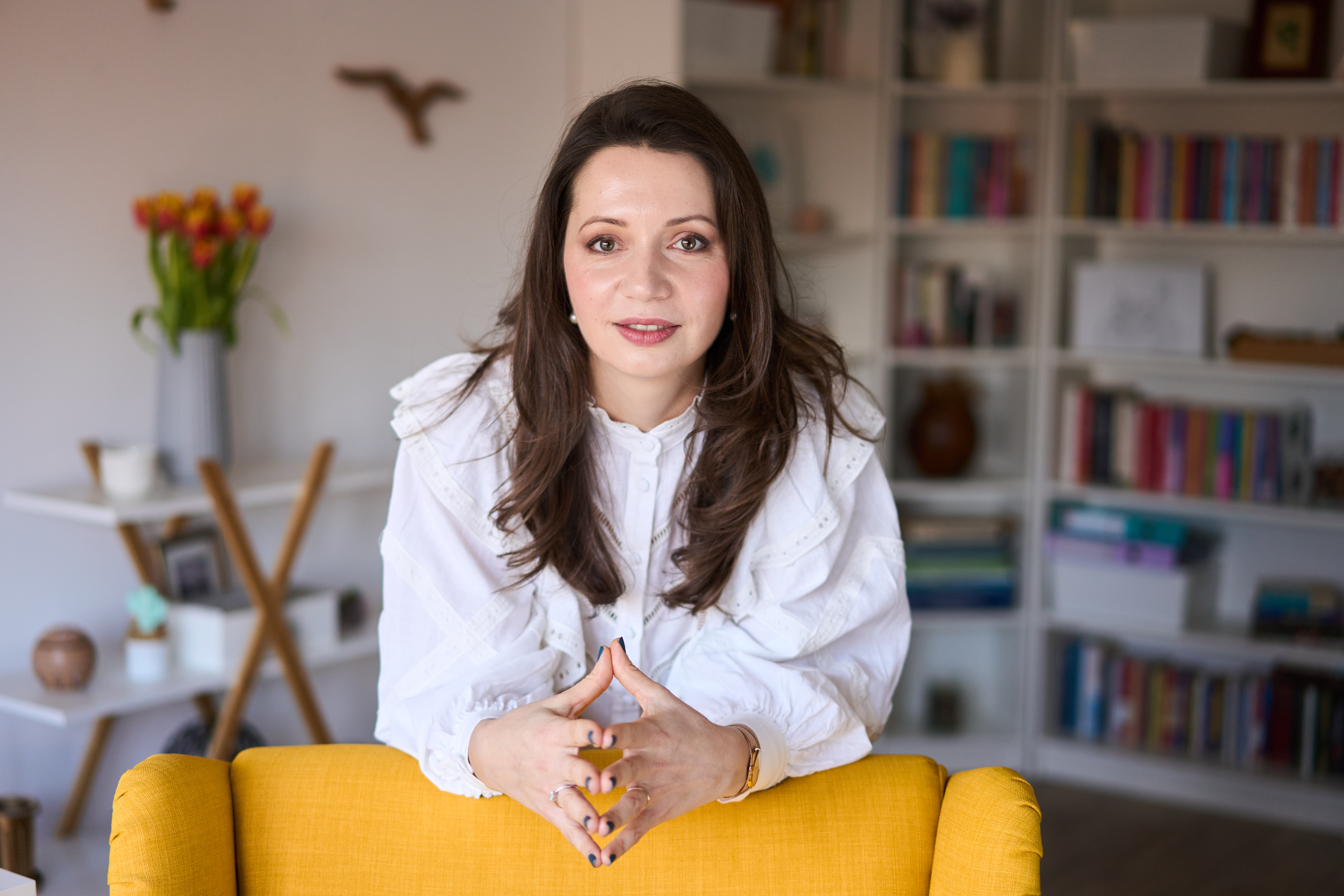 Livia Gheorghe, psihoterapeut, așezată în fotoliul galben, cu biblioteca în fundal