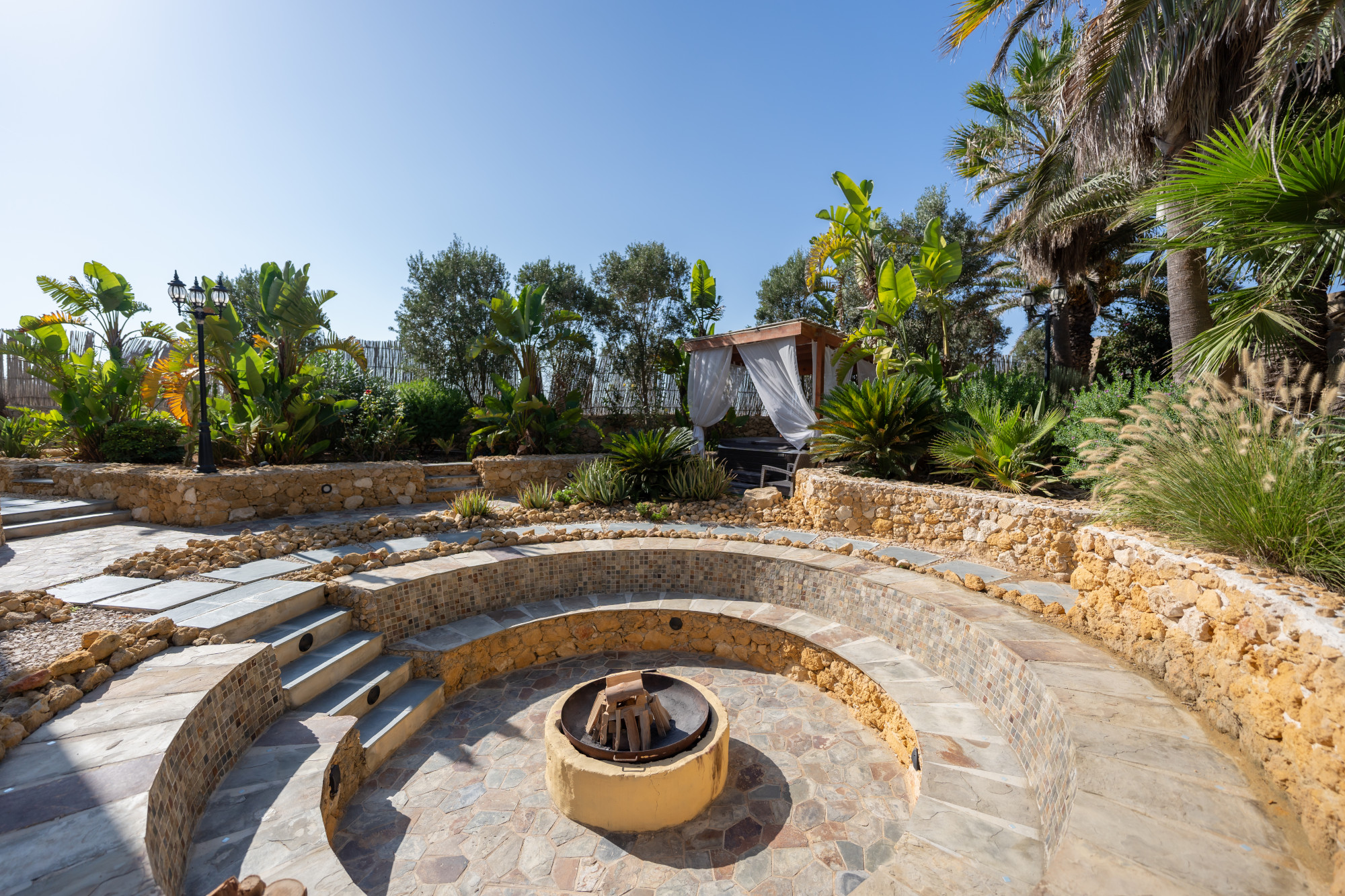 Stone amphitheatre fire pit lounge surrounded by palm trees and Mediterranean gardens
